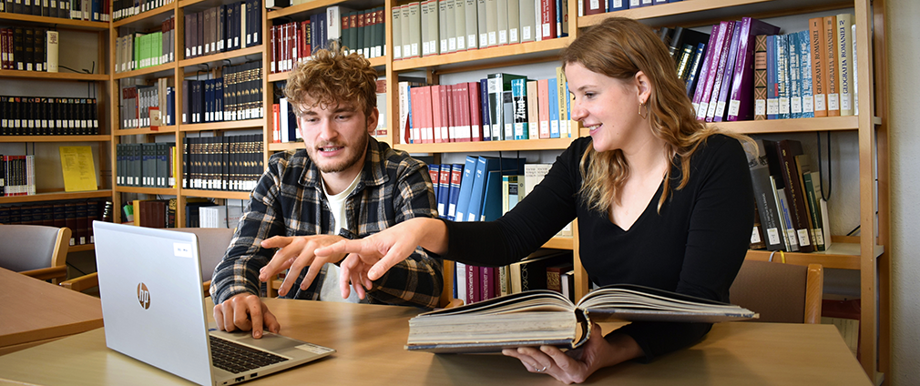 Eine Studentin und ein Student sind in der Bibliothek und schauen gemeinsam auf den Bildschirm eines Notebooks.